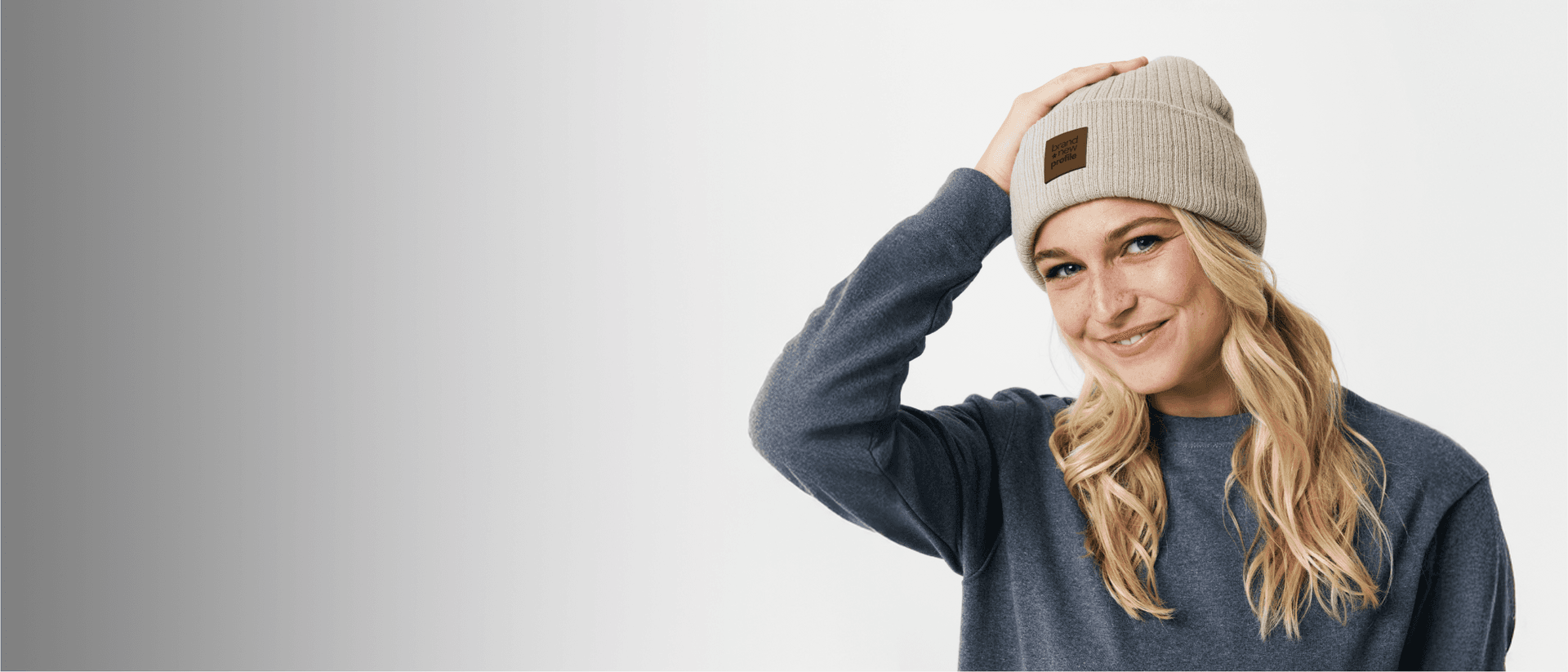 Leende kvinna som justerar en beige, ribbstickad mössa med ett lädermärke som har en egen logotyp.